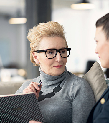 &Quot;In This Image, A Woman Accountant Is Wearing Black-Framed Glasses While Speaking With A Tax Client Seeking Our Taxation Services. She Holds A Pen In Her Right Hand And A Booklet In Her Left Hand, Ready To Take Notes. Both Individuals Are Engaged In Conversation And Appear Focused On The Discussion At Hand.&Quot;